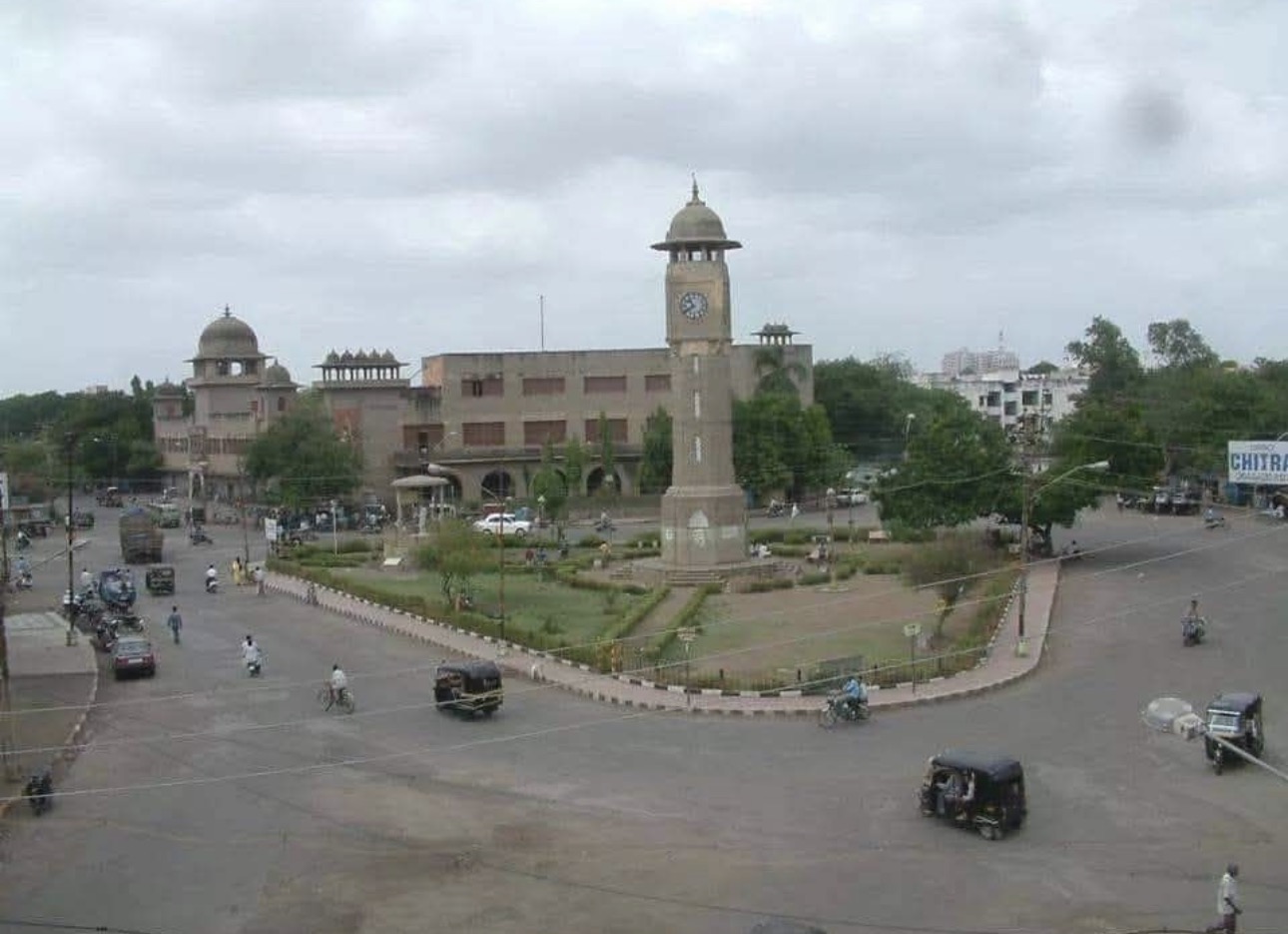 A tall clock tower stands beside a domed old building over a wide square with a round garden, auto-rickshaws and scooters on the road. Colour.
