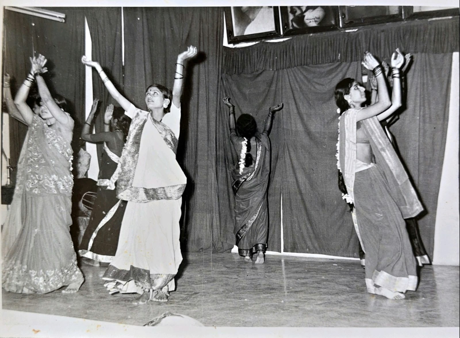 Dancers on stage, arms raised in a single movement. Some wear saris and some wear ghagra-odhni with jasmine in their hair. One dancer at the centre faces away from the audience towards the backdrop. Framed portraits hang above the stage curtain. Black-and-white. Undated. Do you recognise anyone, or the hall?
