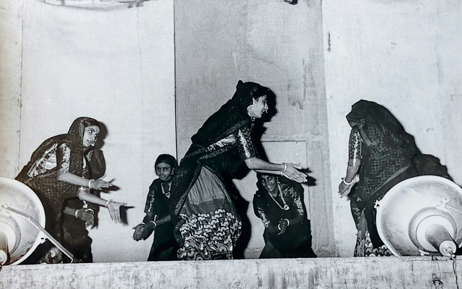 Women and young girls dancing across a stage, bending low with arms swept out, large white props at the edges. Black-and-white.