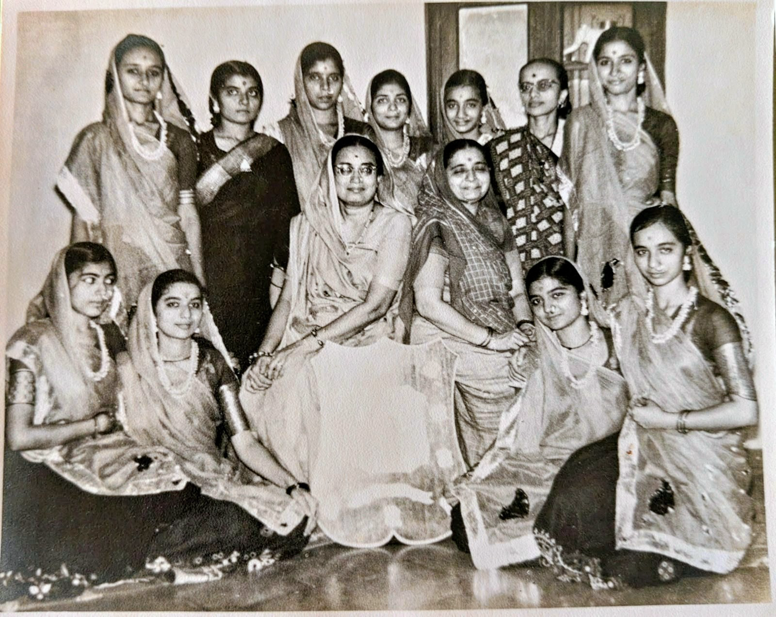 Pragnaben Oza (seated, right, in a checked sari with the end over her head) with the young dancers of her group, among them three of her daughters. Standing directly behind her, smiling, is Poyani Oza. In the front row, second from left, is Yaminiben Oza, and at the far right is Shobhanaben Oza (later Mehta), both Poyani's elder sisters. Probably Nupur Zankar, 1960s. Do you recognise anyone else, including the other seated woman?