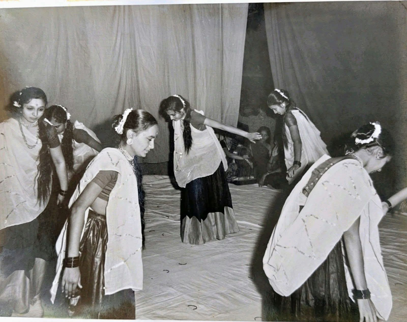 Poyani Oza (centre, bending forward with one arm stretched out, in a dark skirt with a light border, a white odhni and jasmine in her hair) dancing garba on stage with her group. Singers and musicians sit on the floor at the back. Undated. Do you recognise the other dancers?
